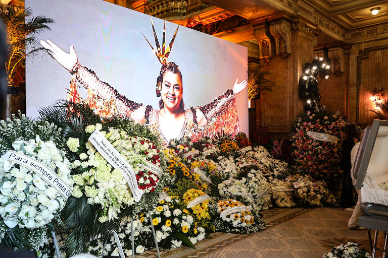 A imagem mostra um grande painel com a foto de uma mulher sorridente, vestida com um traje colorido e adornos. Na frente do painel, há uma grande quantidade de arranjos de flores brancas e coloridas, com algumas faixas que contêm mensagens de homenagem. O ambiente parece ser um local de velório ou homenagem, com uma decoração elaborada e um clima solene.