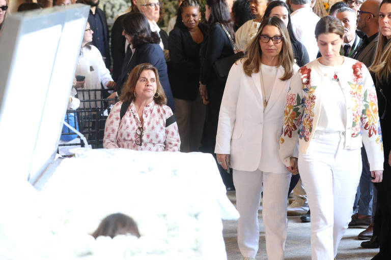 A imagem mostra um grupo de pessoas em uma cerimônia de luto. No primeiro plano, duas mulheres vestindo roupas brancas caminham em direção a um caixão coberto com flores. Uma mulher à esquerda, com uma blusa de estampas, observa a cena. Ao fundo, há várias pessoas, algumas olhando para o caixão, em um ambiente que parece ser um salão de velório.