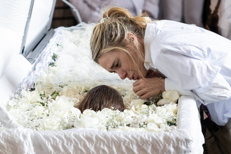Uma mulher está inclinada sobre um caixão branco, que está decorado com flores brancas. Ela parece estar em um momento de tristeza e reflexão, olhando para a pessoa dentro do caixão. O ambiente ao redor é sutil, com outras pessoas ao fundo, mas não são claramente visíveis.