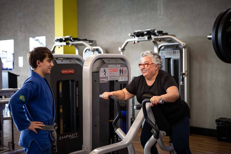 The image shows a boy and a woman in a gym. The boy, dressed in a blue uniform, stands next to a piece of weight training equipment, while the woman, with gray hair and glasses, sits on an exercise machine, smiling and interacting with the boy. In the background, there are other pieces of weight training equipment and a gray wall.