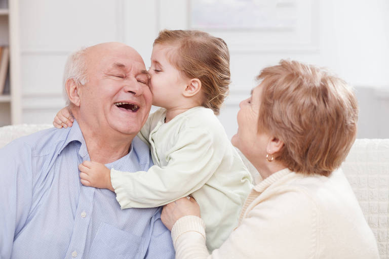 The image shows a smiling grandfather being kissed on the cheek by a small girl, while an older woman, presumably the grandmother, looks on with a smile. The setting is bright and welcoming, with a sofa and shelves in the background.