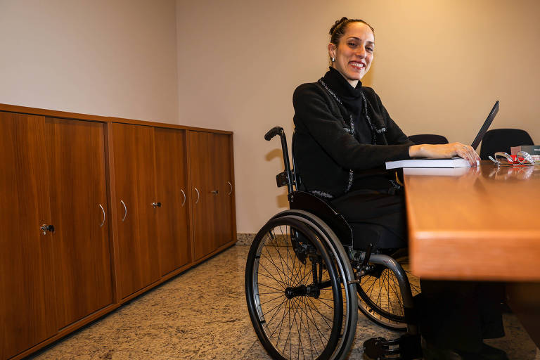 Uma mulher sentada em uma cadeira de rodas, sorrindo, enquanto trabalha em um laptop em uma mesa de escritório. Ela está vestindo uma blusa preta e tem cabelo preso. Ao fundo, há armários de madeira e uma parede clara.