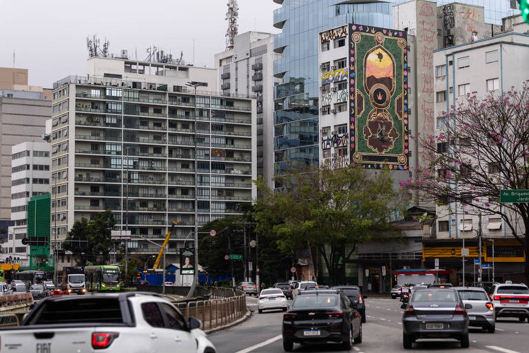 A imagem mostra uma vista urbana de um trecho da cidade, com prédios de diferentes alturas e estilos arquitetônicos. À esquerda, há um edifício residencial de vários andares, enquanto à direita se destaca um mural colorido em uma das fachadas dos prédios. No primeiro plano, há uma via com vários carros em movimento e árvores com flores roxas. O céu está nublado, sugerindo um dia cinzento.