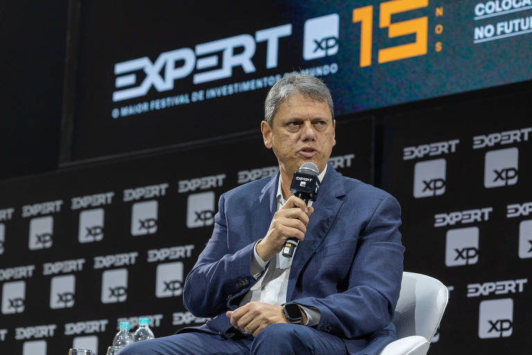 A man sits on a stage, holding a microphone and speaking. He is dressed in a blue suit and a light-colored shirt. In the background, there is a panel with the 'EXPERT' logo and the phrase 'THE WORLD'S LARGEST INVESTMENT FESTIVAL.' The setting appears to be a conference event, with several black signs bearing the 'EXPERT' logo.