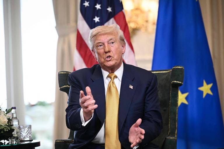 Um homem de cabelo loiro e pele clara está sentado em uma cadeira, gesticulando com as mãos enquanto fala. Ele usa um terno escuro com uma gravata amarela. Ao fundo, há uma bandeira dos Estados Unidos e uma bandeira da União Europeia. Uma mesa com um vaso de flores está visível à esquerda.