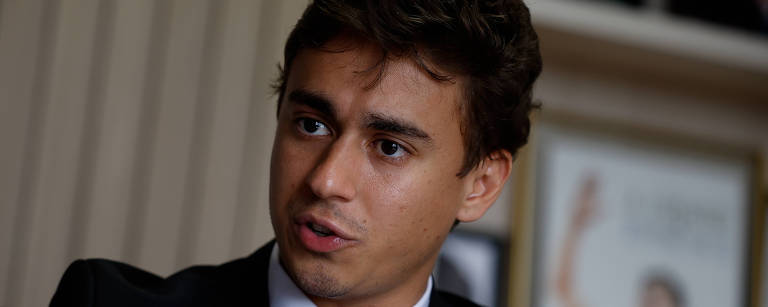 Um jovem homem está sentado em uma cadeira, vestindo um terno escuro e uma gravata azul. Ele está gesticulando com a mão esquerda enquanto fala, com uma expressão séria. Ao fundo, há uma parede com quadros e documentos, sugerindo um ambiente de escritório ou reunião.