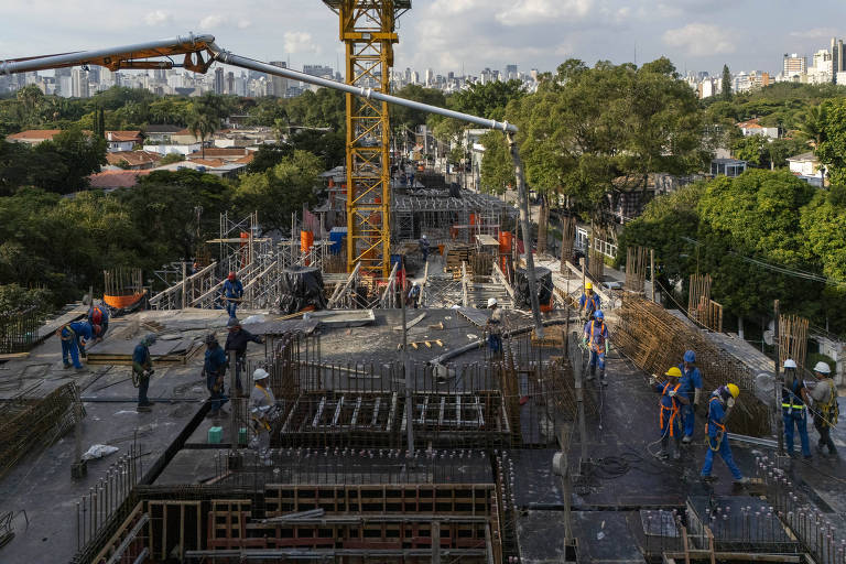 A imagem mostra um canteiro de obras em um edifício em construção. Vários trabalhadores estão presentes, utilizando equipamentos de segurança, como capacetes e coletes. Há uma grua amarela ao fundo, e a estrutura do edifício está em fase de concretagem. A área ao redor é verde, com árvores visíveis, e ao fundo, a cidade se estende com prédios e casas.