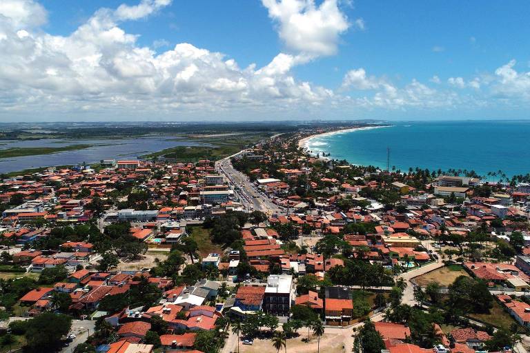 A imagem mostra uma vista aérea de uma cidade litorânea, com casas de telhados vermelhos e ruas visíveis. À esquerda, há uma área verde com água, possivelmente um mangue ou lagoa. À direita, a costa do mar é visível, com ondas e uma praia. O céu está claro, com algumas nuvens brancas.