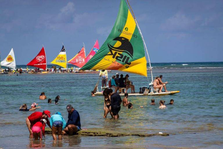 A imagem mostra uma praia com várias pessoas se divertindo na água e na areia. No primeiro plano, algumas crianças estão brincando na água rasa, enquanto um grupo de adultos observa. Ao fundo, há várias embarcações à vela com velas coloridas, incluindo uma com as cores da bandeira do Brasil. O céu está parcialmente nublado e o mar é calmo.