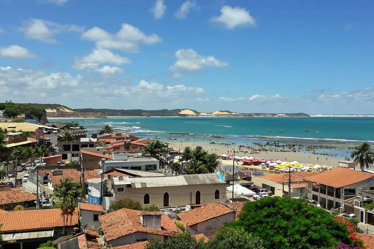 A imagem mostra uma vista panorâmica de uma praia com águas claras e calmas. No primeiro plano, há várias casas com telhados de cerâmica e algumas palmeiras. Ao fundo, é possível ver a areia da praia e algumas barracas de praia. O céu está limpo com algumas nuvens brancas e há formações de dunas ao longe.