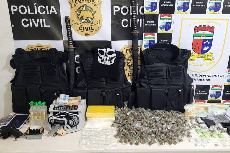 A imagem mostra uma mesa com diversos itens apreendidos pela polícia. Há três coletes à prova de balas pretos, duas armas longas, e uma variedade de drogas, incluindo pacotes de maconha e outros materiais. Também estão presentes frascos, balanças, e celulares, todos dispostos sobre a mesa. Ao fundo, há um painel com logotipos da Polícia Civil e de outras instituições.