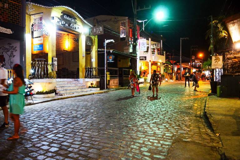 A imagem mostra uma rua pavimentada com pedras, iluminada por luzes artificiais durante a noite. À esquerda, há estabelecimentos comerciais com letreiros visíveis, enquanto à direita, algumas pessoas caminham pela calçada. A atmosfera é vibrante, com várias luzes coloridas ao fundo.