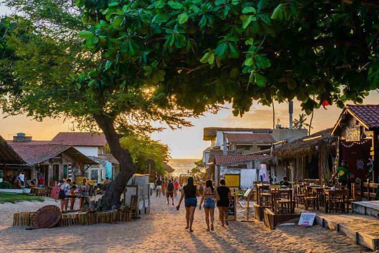 A imagem mostra um caminho de areia em uma praia, cercado por árvores e construções. Há pessoas caminhando pelo caminho, algumas em grupo e outras sozinhas. Ao fundo, é possível ver o mar e o céu com tons de laranja e amarelo, indicando o entardecer. As construções à esquerda e à direita são de estilo rústico, com mesas e cadeiras ao ar livre, sugerindo um ambiente de lazer.