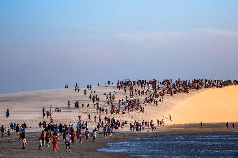 A imagem mostra uma grande duna de areia com muitas pessoas subindo e descendo. No primeiro plano, há um grupo de pessoas caminhando na areia e na água, enquanto no topo da duna, há uma multidão de pessoas. O céu é claro, com tons de azul e algumas nuvens. A cena transmite uma sensação de atividade e lazer ao ar livre.