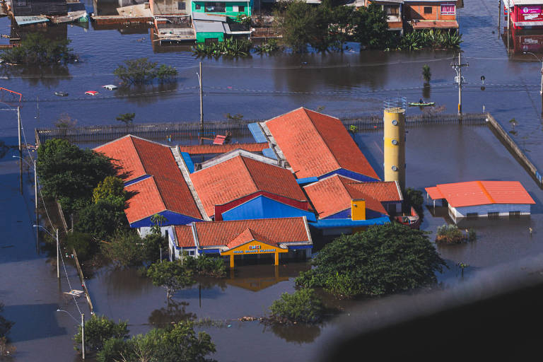 A imagem mostra uma área urbana inundada, com várias casas e edifícios parcialmente submersos em água. Um edifício com telhado vermelho e azul é visível no centro, cercado por água. Ao fundo, há outras construções e vegetação, também afetadas pela inundação. A cena sugere um impacto significativo de chuvas ou enchentes na região.