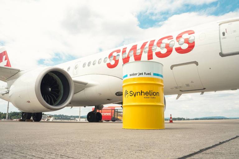 A imagem mostra um avião da companhia aérea Swiss, com a fuselagem branca e a marca em vermelho. Ao lado do avião, há um barril amarelo com a inscrição 'Synthetik'. O fundo apresenta um céu parcialmente nublado e a pista do aeroporto.