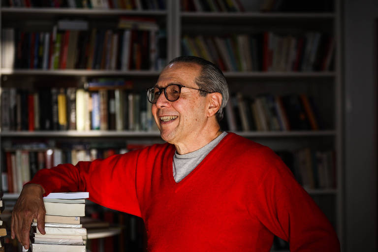 Um homem está sorrindo enquanto se apoia em uma pilha de livros. Ele usa um suéter vermelho e óculos. Ao fundo, há uma estante cheia de livros. A luz do sol ilumina parcialmente a cena, criando um contraste entre luz e sombra.