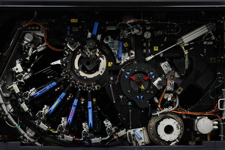 A imagem mostra o interior de uma máquina complexa, com diversos componentes mecânicos e eletrônicos visíveis. Há um grande disco central com braços e engrenagens, além de fios e conectores coloridos que se espalham por toda a estrutura. O fundo é escuro, destacando os detalhes da máquina.