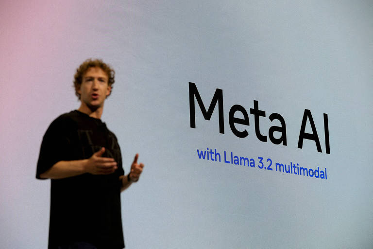 Um homem está em pé, gesticulando enquanto fala em uma apresentação. Ao fundo, há um grande texto que diz 'Meta AI' em letras grandes e em negrito, com a frase 'with Llama 3.2 multimodal' logo abaixo em letras menores. O homem usa uma camiseta preta e tem cabelo cacheado e loiro.