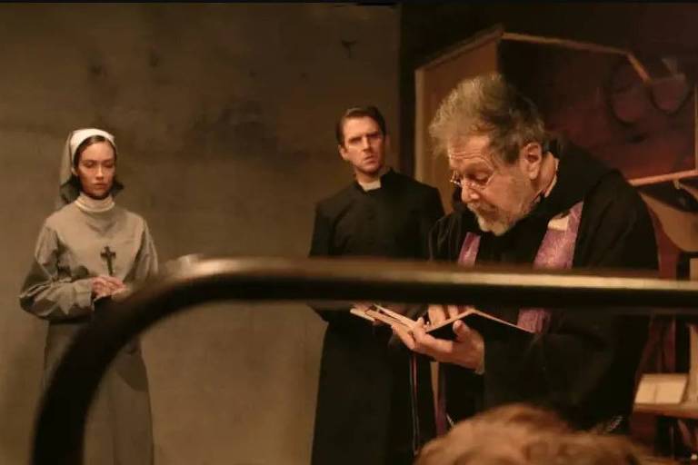 A imagem mostra um grupo de quatro pessoas em um ambiente interno, possivelmente uma sala ou um espaço de culto. À esquerda, duas mulheres vestidas com hábitos religiosos estão de pé, segurando livros. Uma delas tem um olhar sério e a outra parece atenta. No centro, um homem vestido de preto, que parece ser um sacerdote, está lendo um texto de um livro, enquanto um outro homem, também vestido de preto, observa. O ambiente é escuro e rústico, com prateleiras ao fundo contendo objetos variados.