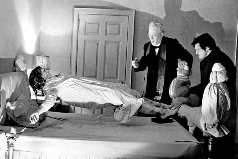 A imagem mostra uma cena dramática em preto e branco, onde uma jovem mulher está levitando sobre uma cama. Ela está vestida com um vestido de noite e parece estar em estado de transe. Ao redor dela, há três homens: um deles, com cabelo grisalho e vestindo um terno escuro, parece estar realizando um ritual, enquanto os outros dois, um com um olhar preocupado e o outro segurando seus pés, observam a situação. O ambiente é sombrio, com luzes e sombras projetadas nas paredes.