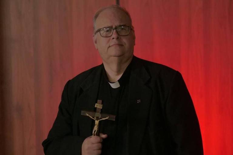 Um homem de meia-idade, vestido com uma batina preta e óculos, segura um crucifixo em uma mão. Ele está em pé em frente a um fundo de madeira com iluminação vermelha, que cria um contraste com sua roupa escura. O homem tem uma expressão séria e parece estar em um ambiente que sugere um contexto religioso.