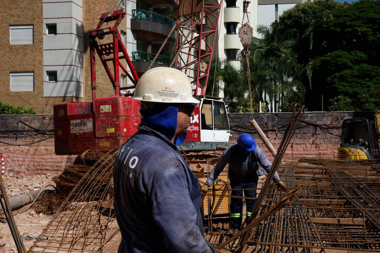 A imagem mostra um canteiro de obras com dois trabalhadores. Um deles, à esquerda, usa um capacete branco e uma camisa azul, enquanto o outro, à direita, está agachado e usa um capacete azul. Ao fundo, há uma grua vermelha e um caminhão branco, além de edifícios e árvores ao redor. O chão está coberto com vergalhões de aço e terra.