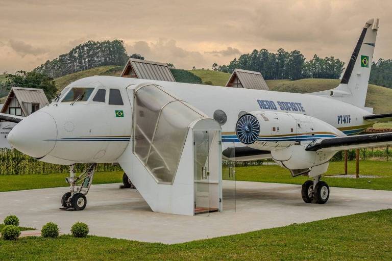 Un piccolo aereo è parcheggiato in un'area aperta circondata da erba verde. L'aereo è bianco con dettagli blu e ha la bandiera brasiliana sulla fusoliera. Accanto all'aereo c'è una struttura in vetro che sembra un ingresso o una cabina. Sullo sfondo si vedono colline ricoperte di vegetazione e alcuni edifici in stile rustico.