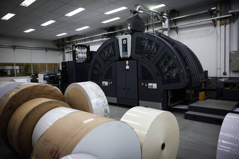 A imagem mostra uma impressora industrial de grande porte em um ambiente de fábrica. À frente da impressora, há rolos de papel em diferentes tamanhos e cores, com alguns rolos sendo brancos e outros em tons de marrom. O ambiente é bem iluminado, com um teto alto e paredes claras, e a impressora possui um painel de controle visível na parte frontal.