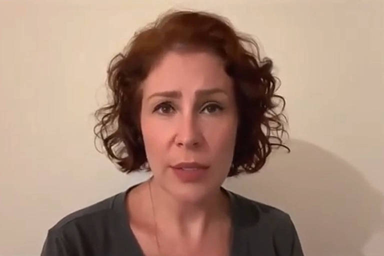 A imagem mostra uma mulher com cabelo cacheado e castanho claro, usando uma camiseta cinza. Ela está olhando diretamente para a câmera com uma expressão séria. O fundo é uma parede clara, sem detalhes visíveis.