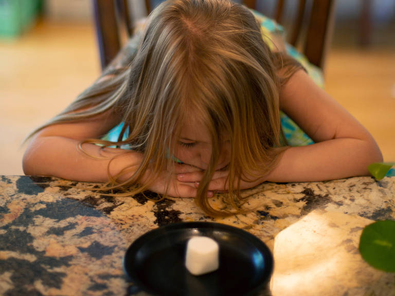 Un niño de pelo largo y liso se inclina sobre una mesa de granito, mirando fijamente un malvavisco blanco sobre un plato negro. La habitación está bien iluminada, con un fondo desenfocado que sugiere una cocina o sala de estar. El niño parece concentrado y curioso.
