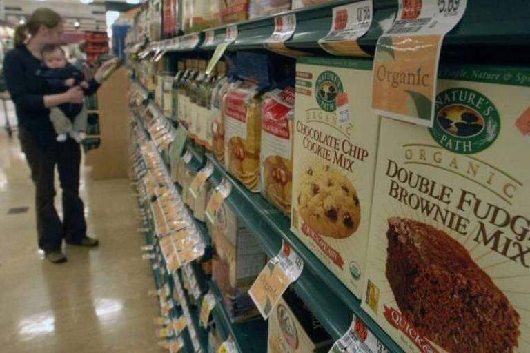 A imagem mostra uma prateleira de supermercado com diversos produtos alimentícios, incluindo misturas para bolos e biscoitos. Uma mulher está segurando um bebê enquanto observa os produtos. Os rótulos dos produtos são visíveis, destacando misturas orgânicas, como 'Double Fudge Brownie Mix' e 'Chocolate Chip Cookie Mix'. O ambiente é bem iluminado e organizado, com prateleiras alinhadas.