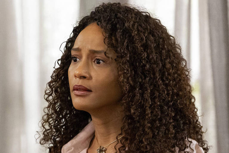 A imagem mostra uma mulher com cabelo cacheado e castanho escuro, usando uma camisa rosa clara. Ela está com uma expressão facial de preocupação ou reflexão, olhando para o lado. Ao fundo, há cortinas claras que filtram a luz, criando um ambiente suave e iluminado.