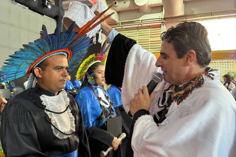 A imagem mostra uma cerimônia cultural com duas pessoas em destaque. À esquerda, um homem com um traje tradicional indígena, incluindo uma grande pluma colorida na cabeça. À direita, um homem vestido com um traje branco e adornos coloridos, segurando um microfone e falando. Ao fundo, outras pessoas em trajes tradicionais estão visíveis, criando um ambiente festivo e cultural.