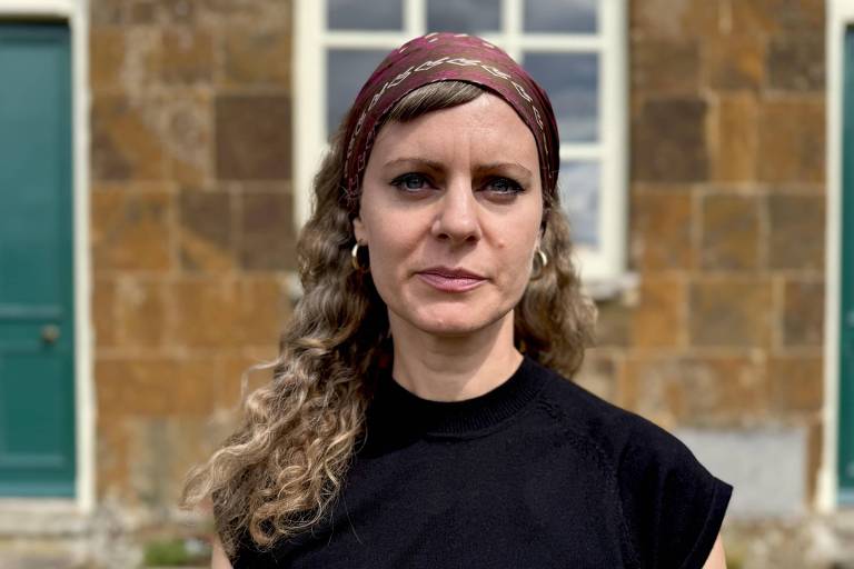A imagem mostra uma mulher com cabelo cacheado e longo, usando uma blusa preta e um lenço na cabeça. Ela está em frente a uma parede de pedra com duas portas verdes ao fundo. A mulher tem uma expressão séria e está olhando diretamente para a câmera.