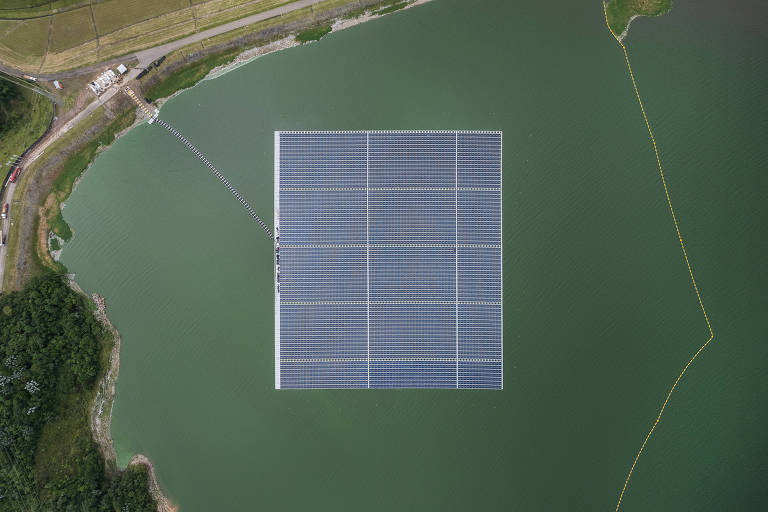 A imagem mostra uma usina solar flutuante instalada sobre um corpo d'água. A estrutura retangular composta por painéis solares está posicionada no centro do lago, cercada por água verde. À esquerda, é possível ver uma área de terra com vegetação e uma estrada. No fundo, há uma pequena construção e uma linha de separação na água.