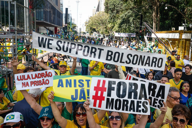 A imagem mostra uma multidão em um protesto, com pessoas segurando cartazes. Os cartazes têm as seguintes inscrições: 'WE SUPPORT BOLSONARO', 'LULA LADRÃO NA PRISÃO', 'ANISTIA JÁ' e '#FORA MORAES'. As pessoas estão vestidas com camisetas nas cores verde e amarelo, típicas do Brasil, e há uma grande quantidade de bandeiras do Brasil ao fundo. O ambiente é urbano, com prédios visíveis.