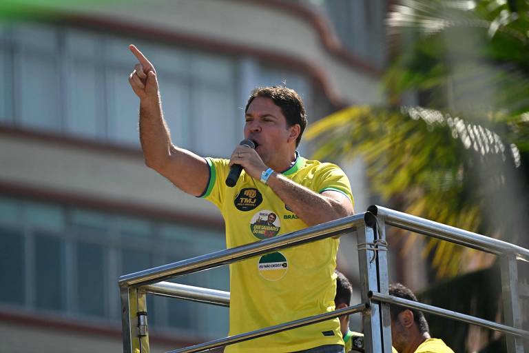 Um homem em uma plataforma elevada está fazendo um discurso. Ele usa uma camiseta amarela e segura um microfone com a mão direita, enquanto aponta com a mão esquerda. Ao fundo, há um edifício e algumas palmeiras, indicando que o evento ocorre ao ar livre.