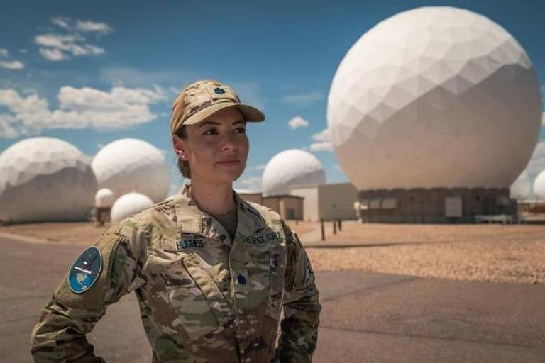Une femme en uniforme militaire se tient dehors, avec plusieurs grandes structures sphériques blanches en arrière-plan. Le ciel est dégagé avec quelques nuages. La femme a les cheveux attachés et porte une casquette. Les sphères semblent être des antennes ou des équipements de communication.