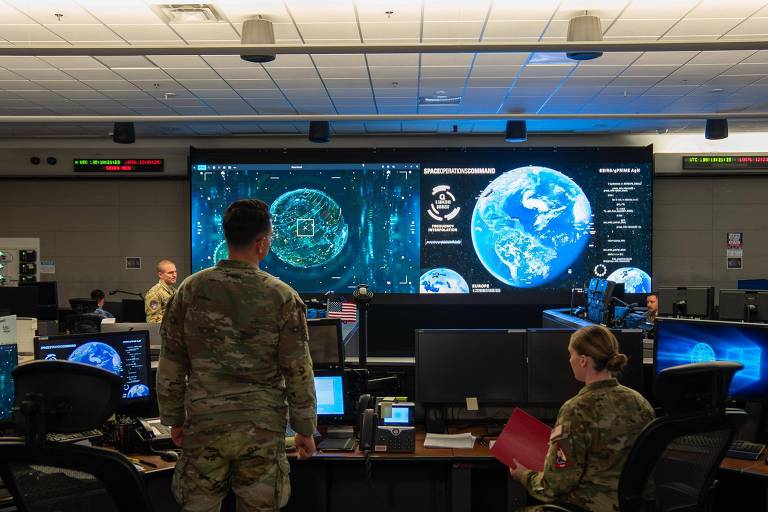 L'image montre un centre de commandement militaire équipé de plusieurs grands écrans diffusant des informations sur l'espace et la Terre. Deux militaires y figurent en bonne place, l'un de dos tourné, l'autre assis, tous deux en tenue de camouflage. L'environnement est moderne, avec plusieurs bureaux et ordinateurs. Les écrans affichent des graphiques et des cartes, indiquant les activités de surveillance.