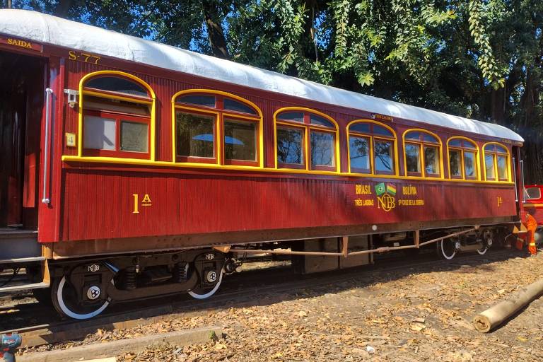 A imagem mostra um vagão de trem antigo, pintado em um tom de vermelho escuro com detalhes em amarelo. O vagão possui janelas grandes e arredondadas, e a parte lateral exibe inscrições em letras douradas. O ambiente ao redor é natural, com árvores ao fundo e um chão de terra e madeira.