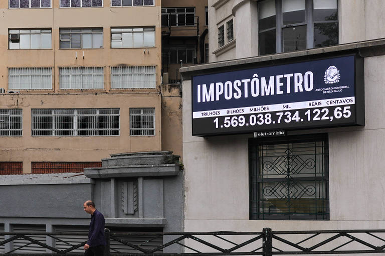 A imagem mostra um painel chamado 'Impostômetro', que exibe um número grande em destaque: 1.569.038.734.122,65. Abaixo, estão as palavras 'TRILHÕES', 'BILHÕES', 'MILHÕES', 'MIL', 'REAIS' e 'CENTAVOS'. O painel está fixado em um edifício, e ao fundo, há outros prédios. Uma pessoa está caminhando na calçada em frente ao painel.
