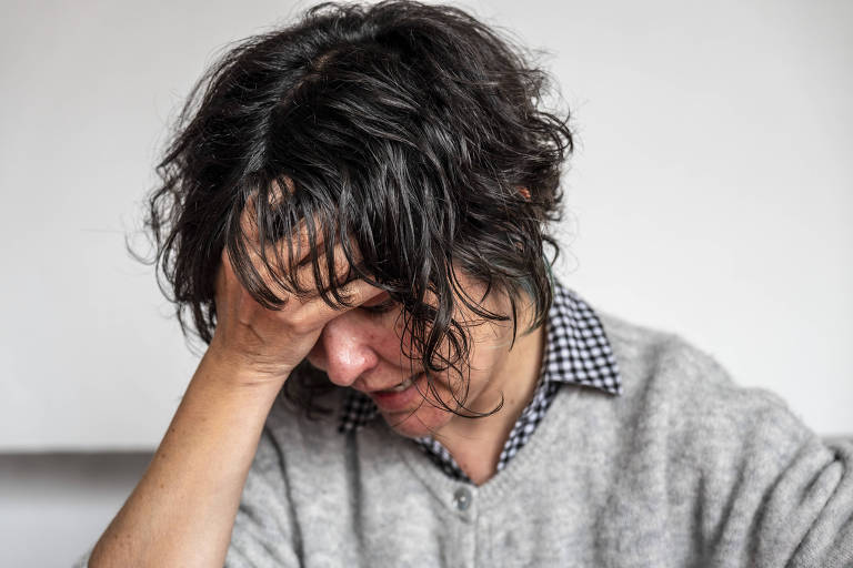 De afbeelding toont een vrouw met donker, krullend haar en een grijze trui. Ze heeft haar hand op haar hoofd, wat bezorgdheid of stress uitdrukt. De achtergrond is neutraal en onscherp, waardoor het figuur van de vrouw wordt benadrukt.