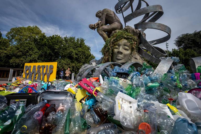 A imagem mostra uma escultura de uma figura humana com um rosto sereno, cercada por uma espiral de metal. Na base da escultura, há uma grande quantidade de lixo plástico, incluindo garrafas e outros objetos coloridos. Um cartaz amarelo com a palavra 'PLASTIC' está em destaque, e ao fundo, duas pessoas estão visíveis, observando a cena. O céu está parcialmente nublado.