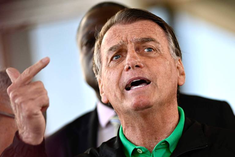 Um homem com cabelo castanho claro e pele clara está falando em um evento. Ele usa uma jaqueta preta com detalhes verdes e gesticula com a mão, apontando para cima. Ao fundo, há uma pessoa com pele escura, parcialmente visível. O ambiente parece ser ao ar livre, com uma iluminação natural.