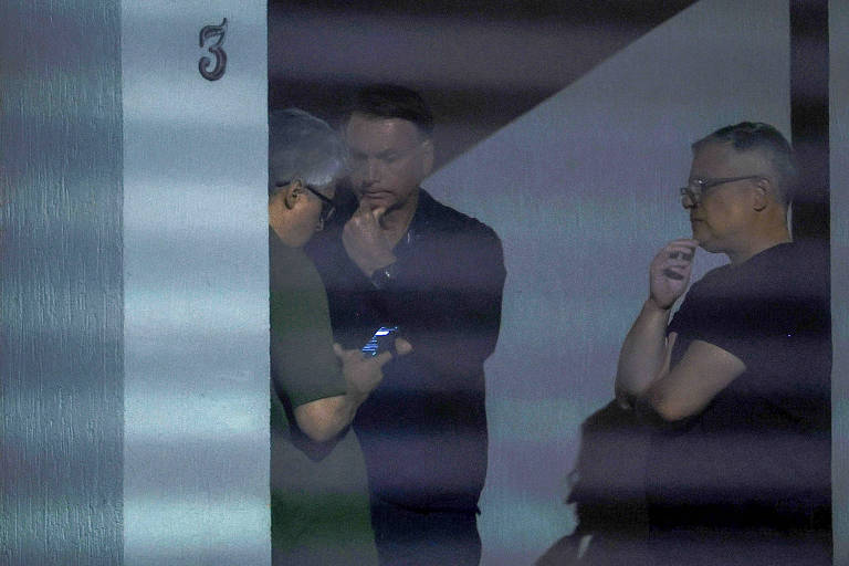 Três homens estão em pé em frente a uma porta numerada com o número 3. Um dos homens, à esquerda, está olhando para um celular. Os outros dois homens estão conversando, um deles com a mão no queixo. A iluminação é suave e há sombras visíveis na parede ao fundo.