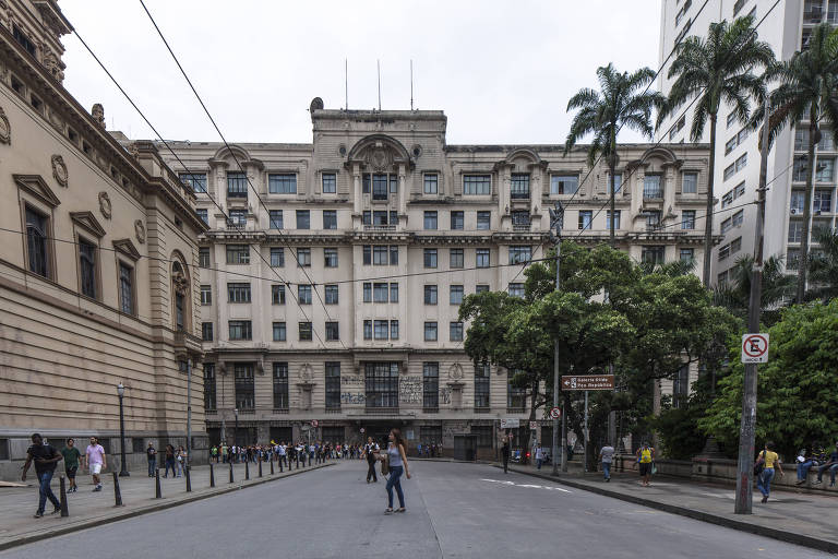 A imagem mostra um edifício de estilo arquitetônico clássico em São Paulo, com uma fachada elaborada e janelas em destaque. À frente do edifício, há uma rua larga com várias pessoas caminhando. À esquerda, pode-se ver uma construção adjacente com detalhes arquitetônicos semelhantes. O céu está nublado e há algumas palmeiras e árvores ao redor, além de placas de sinalização visíveis.