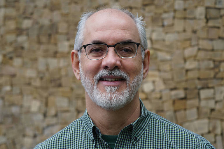 Um homem de meia-idade com cabelo grisalho e barba, usando óculos e uma camisa xadrez verde. Ele está sorrindo e posando para a foto, com um fundo de pedras empilhadas.