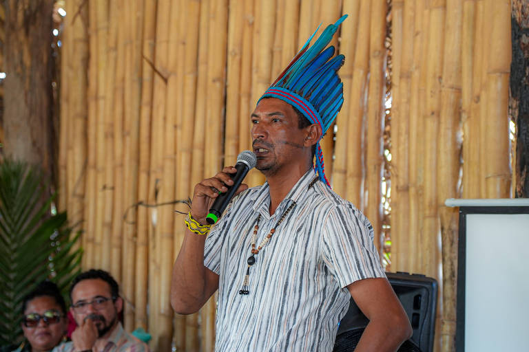Um homem com um cocar colorido e roupas tradicionais está falando em um microfone. Ele está em um ambiente com paredes de bambu e há pessoas sentadas ao fundo, ouvindo atentamente. O homem parece estar em uma palestra ou apresentação.