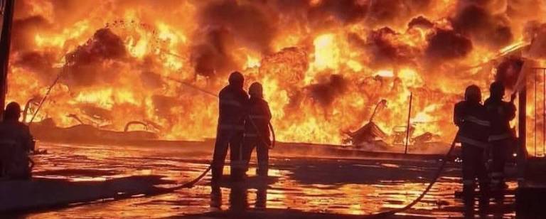 A imagem mostra um incêndio intenso, com chamas e fumaça predominando em tons de laranja e vermelho. Duas silhuetas de pessoas estão em primeiro plano, observando o fogo. O cenário parece ser urbano, com estruturas visíveis ao fundo, mas a visibilidade é limitada devido à fumaça. A atmosfera é dramática e tensa, refletindo a gravidade da situação.
